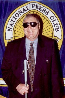 Picture of John Fales - Sgt. Shaft standing in front of a National Press Club flag. This picture is also a link to his column